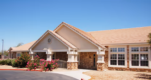 Exterior view of a single-story building with a beige stucco finish and stone accents, featuring a tiled roof and multiple windows. There is a covered entrance with a small ramp and flowering bushes near the sidewalk under a clear blue sky.