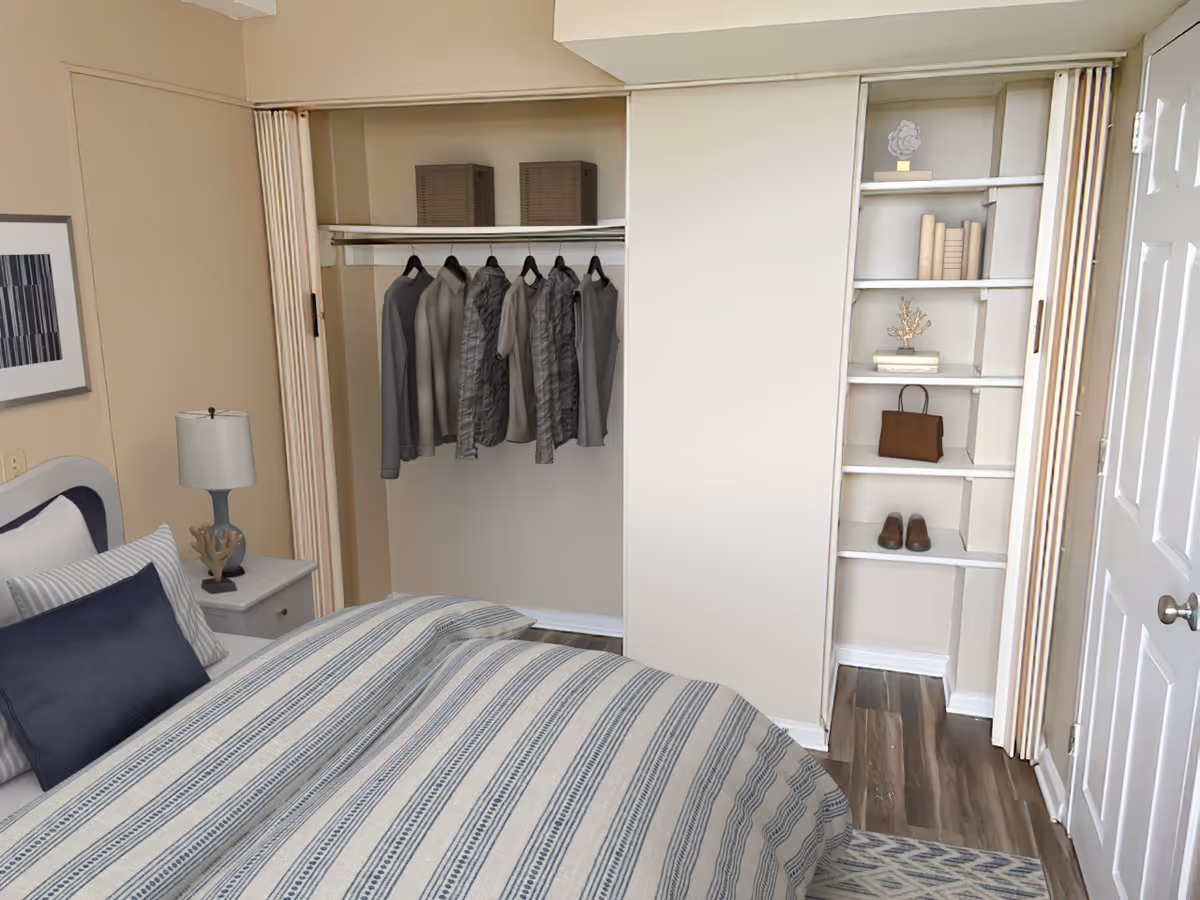 A bedroom with a bed covered in a striped comforter and pillows, a nightstand with a lamp and decorative item, an open closet with hanging clothes and storage baskets, and built-in shelves holding books, a handbag, and shoes.