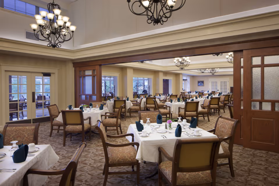 A spacious dining room with multiple tables covered in white tablecloths, each set with plates, cups, silverware, and dark green folded napkins. The room features patterned carpet, upholstered chairs, large windows, and elegant chandeliers hanging from the ceiling. There is a wooden partition with glass panels dividing the space.
