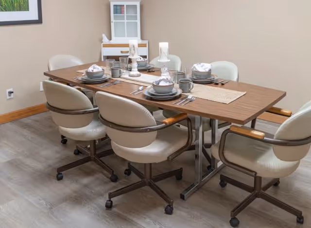 A dining table set for six with rolling chairs and place settings in a neutral-colored room.