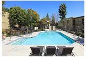 Outdoor rectangular swimming pool with lounge chairs and apartment buildings around it under a clear blue sky.
