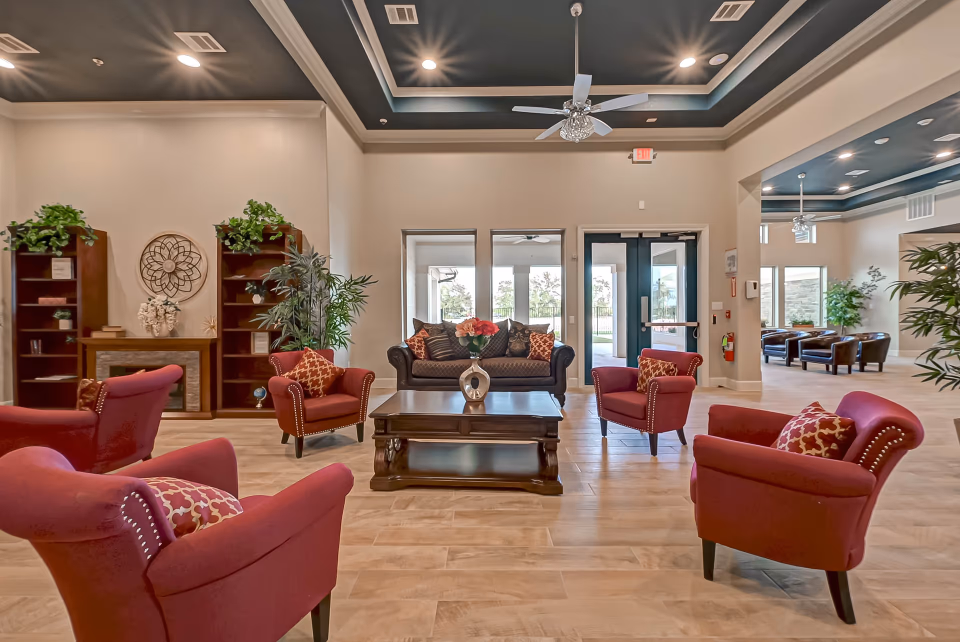 A spacious and well-lit living room area in an assisted living facility featuring a dark brown sofa with patterned cushions, four red armchairs with matching pillows arranged around a wooden coffee table with a decorative vase of flowers. The room has light wood flooring, beige walls, a dark tray ceiling with recessed lighting and ceiling fans, and large windows and glass doors leading to an outdoor area. There are bookshelves with plants and decorative items on the left side and additional seating visible in an adjacent room.