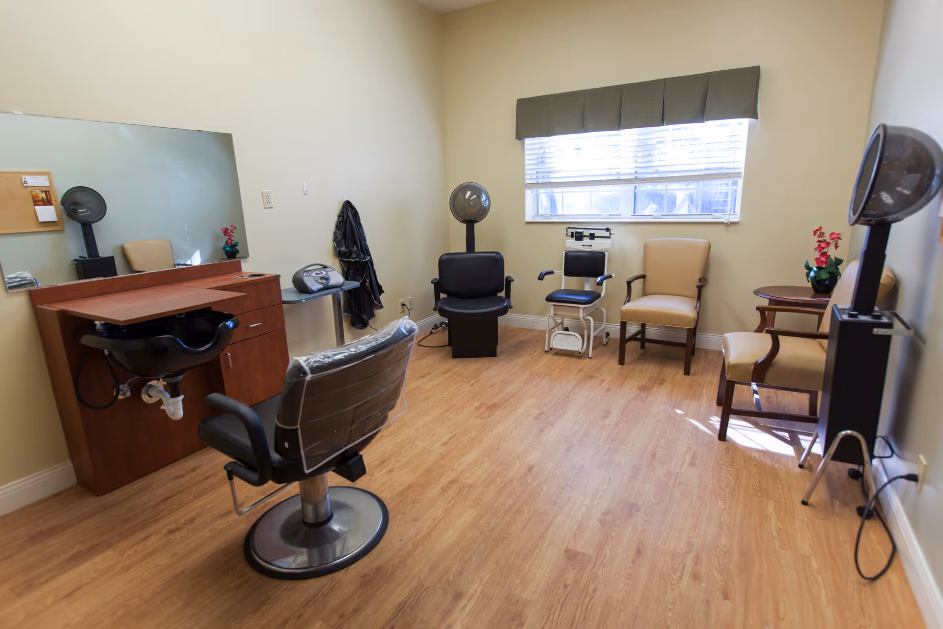 A small salon room with a hair washing station, a styling chair covered in plastic, a large mirror, a hair dryer chair, a scale, and several beige chairs arranged along the walls. The room has light-colored walls, a window with blinds, and wood flooring.