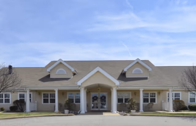 Front exterior view of a single-story senior living facility building with beige siding, white trim, and a covered entrance supported by white columns. The building has two dormer windows on the roof and several windows along the front. There are small shrubs and outdoor chairs near the entrance, and the sky is clear with some light clouds.