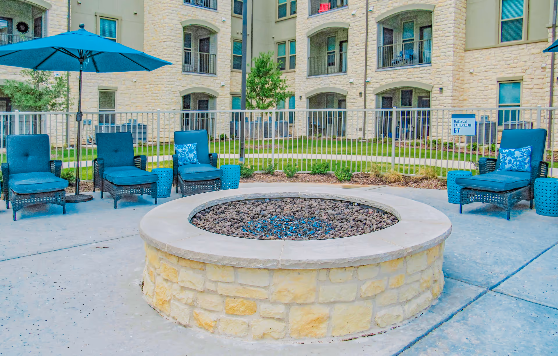 Outdoor courtyard featuring a circular stone fire pit surrounded by teal cushioned lounge chairs and an umbrella in front of an apartment building.