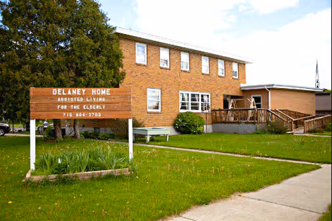 Front view of a two-story brick assisted-living building with a lawn, ramped entrance, and a wooden sign reading 'Delaney Home Assisted Living For The Elderly'.