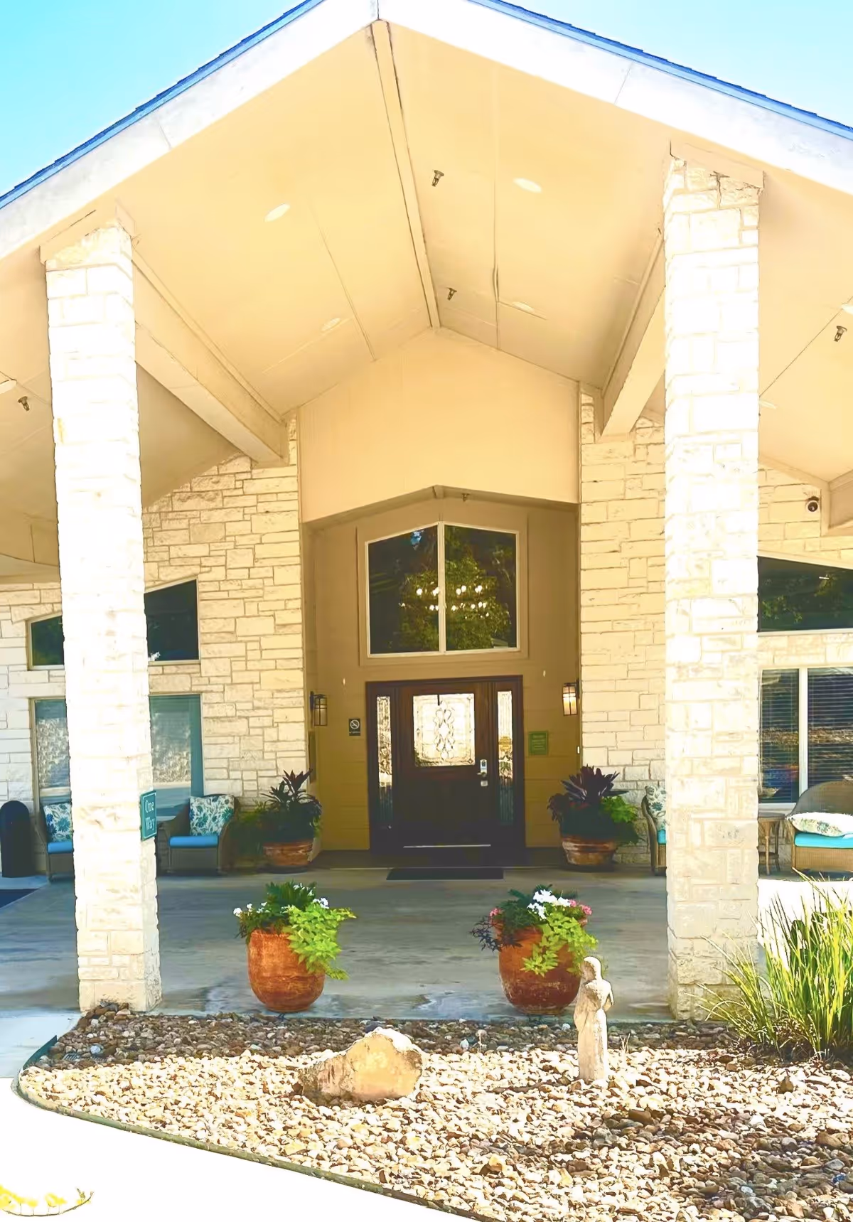 Entrance of Heritage Place of Boerne featuring a covered porch with stone pillars, potted plants, a glass-paneled wooden door, and seating areas on either side.