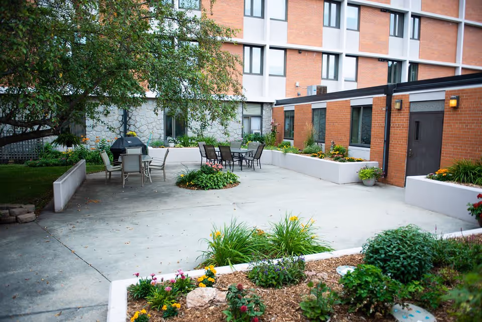 A paved courtyard with outdoor tables and chairs, planters and flower beds beside a multi-story brick building.
