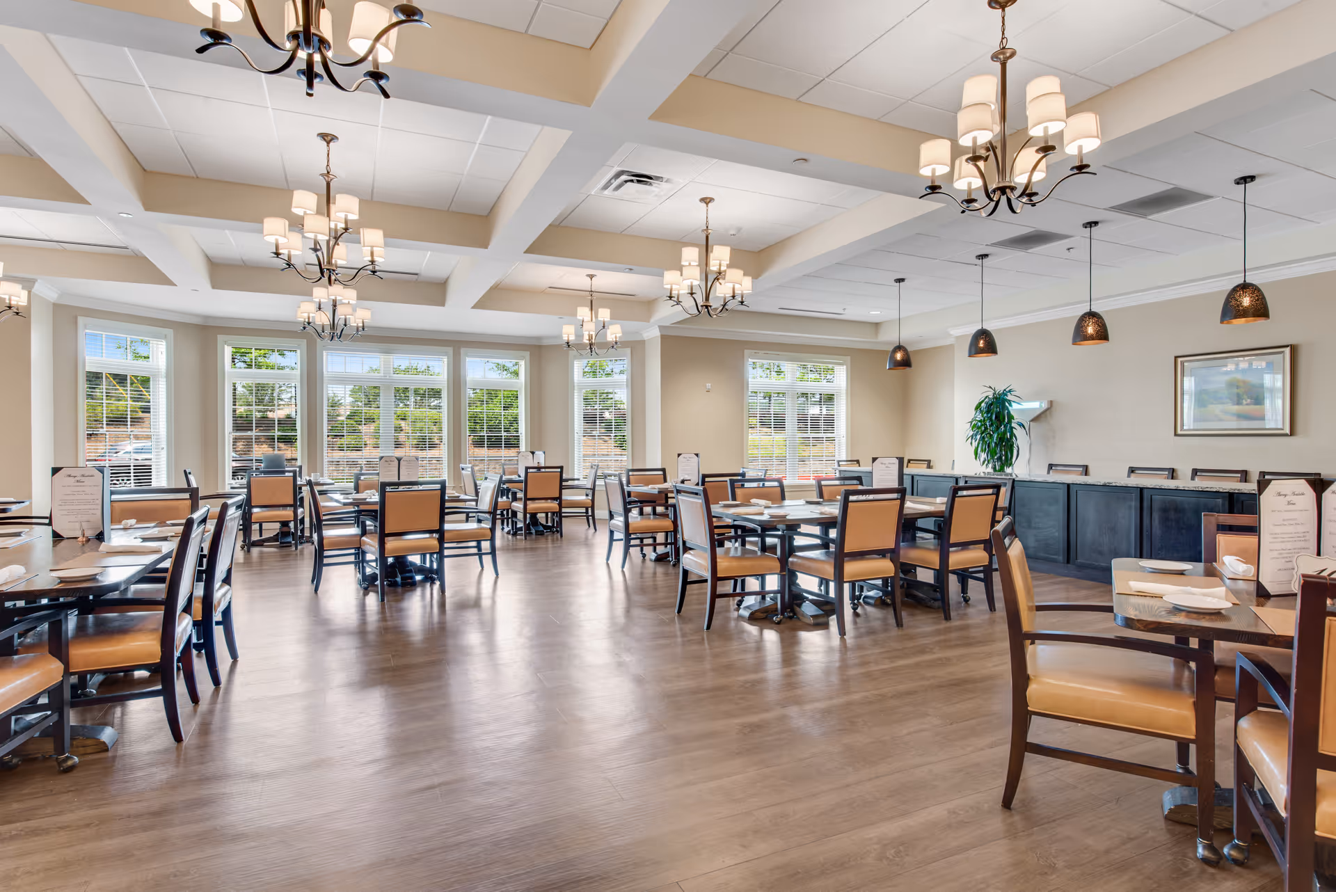 Bright, spacious dining room with multiple tables and chairs, chandeliers, pendant lights, large windows, and wood flooring.