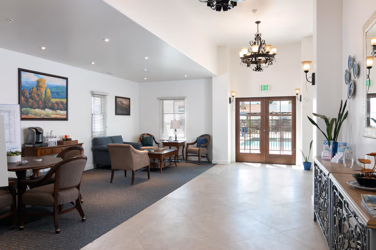 A bright and spacious common area with seating including a blue sofa, armchairs, and wooden tables. There is a coffee station on a wooden sideboard, artwork on the walls, and large glass doors leading outside to a pool area. The room features tiled flooring, carpeted seating area, and decorative light fixtures.
