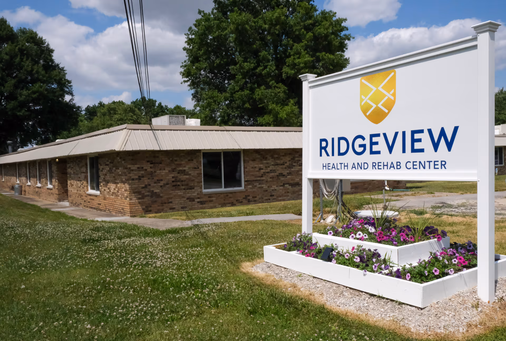 Exterior view of Ridgeview Health and Rehab Center showing a single-story brick building with a metal roof, a large white sign with the facility's name and logo, and a flower bed with purple and white flowers in front of the sign. Trees and a partly cloudy sky are visible in the background.