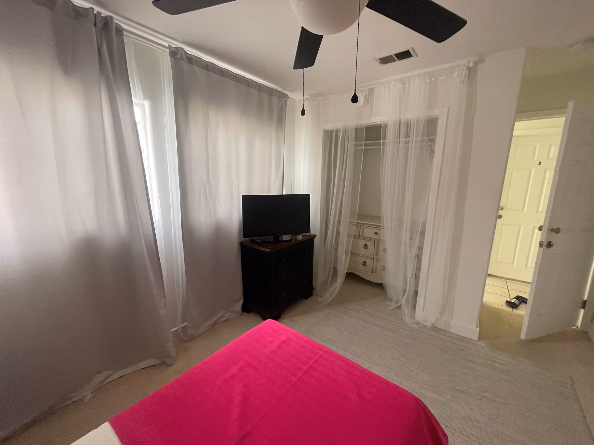 A bedroom with a bed covered in a bright pink blanket in the foreground. There are two windows with light gray curtains on the left wall. A black dresser with a flat-screen TV on top is positioned against the wall. An open closet with sheer white curtains reveals a white dresser inside. A ceiling fan with black blades hangs from the ceiling. The door to the room is open, showing a hallway with tiled flooring and another door.