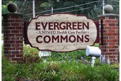 A wooden entrance sign reading 'Evergreen Commons — a NYMED Health Care Facility' mounted between brick pillars with grass and trees in the background.