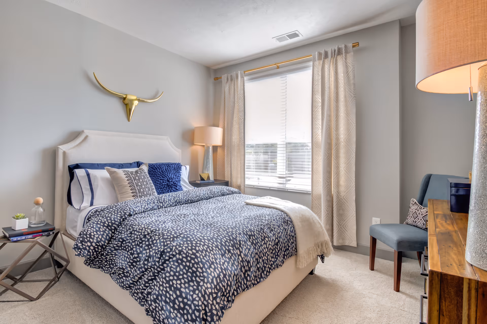 A cozy bedroom with a beige upholstered bed featuring blue and white bedding and decorative pillows. A gold bull skull wall decoration hangs above the bed. There is a bedside table with a lamp and books on the left side, and a wooden dresser with a lamp and a blue chair on the right side. A window with beige curtains and white blinds lets in natural light.