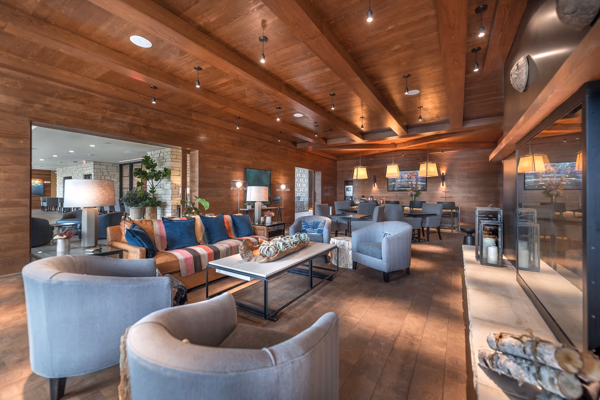 A cozy living room area with wooden walls and ceiling, featuring a brown sofa with colorful cushions, two gray armchairs, a coffee table with decorative plants, and a fireplace with candles and stacked firewood. In the background, there is a dining area with tables, chairs, and hanging pendant lights.