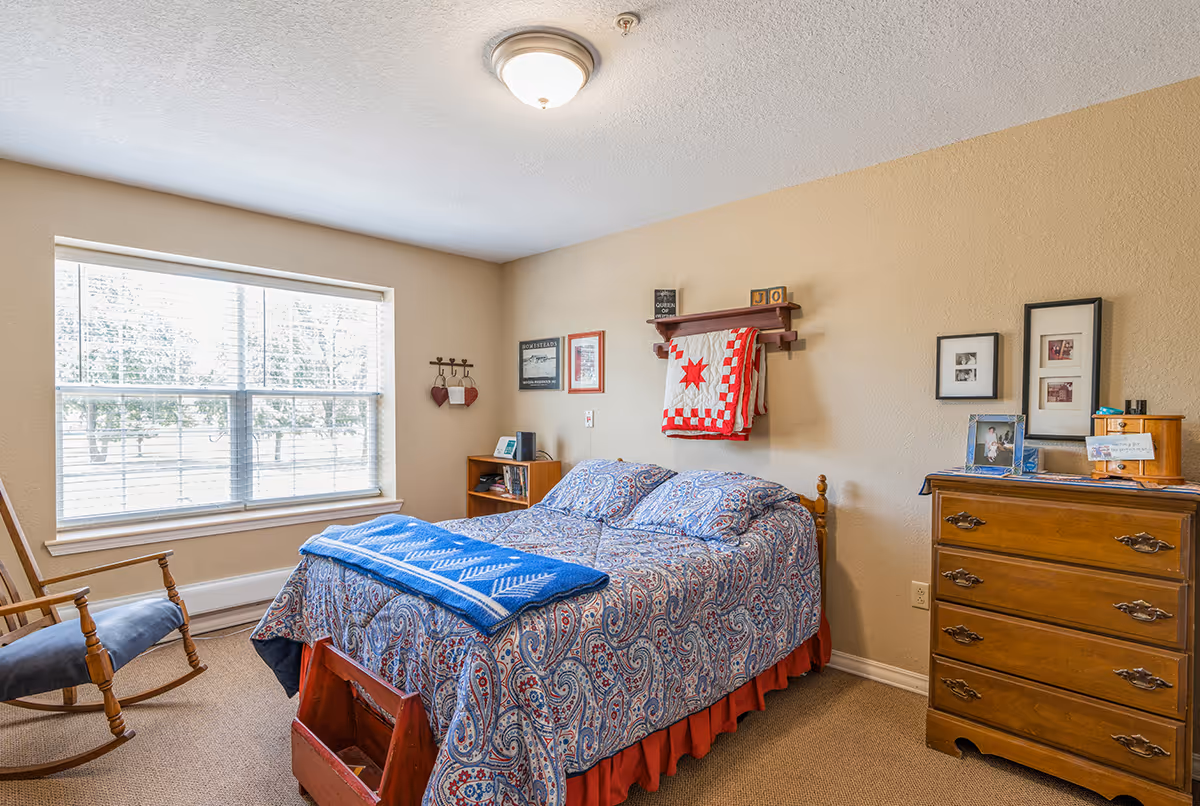A cozy senior living bedroom with a double bed covered in a blue and red patterned bedspread and a blue throw blanket. There is a wooden rocking chair near a large window with blinds, a wooden dresser with framed photos, and a small wooden shelf on the wall holding a red and white quilt and decorative items. The walls are beige, and the room is carpeted.
