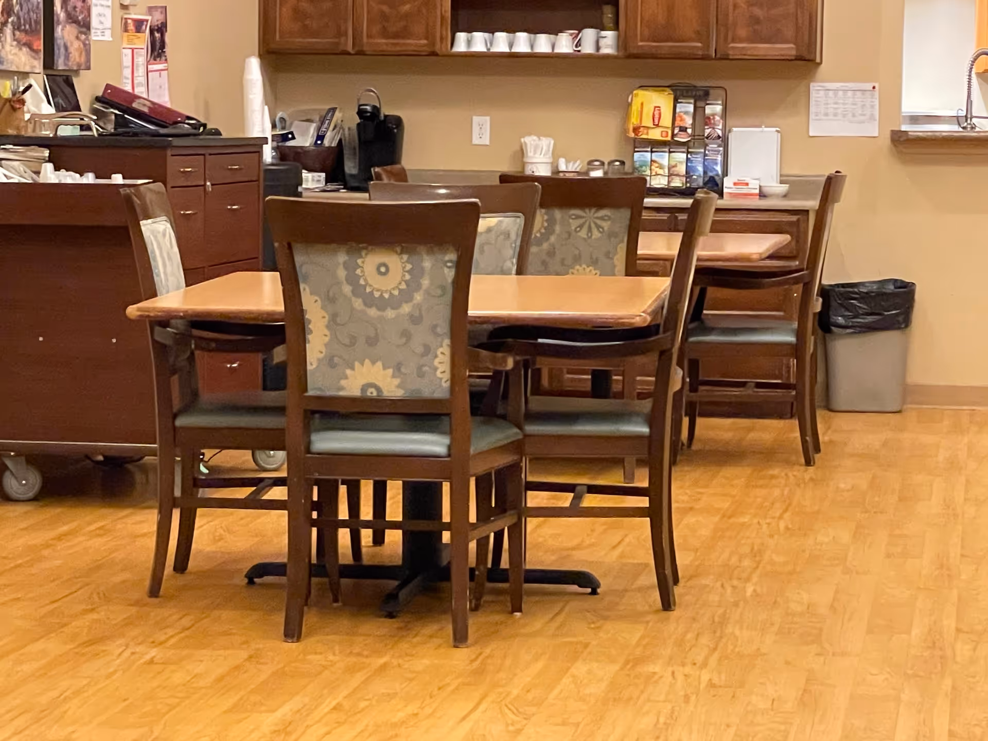 Interior view of a dining area in a senior care facility with wooden tables and chairs featuring floral patterned upholstery. In the background, there are cabinets, a coffee station, and a trash bin against beige walls and wood flooring.