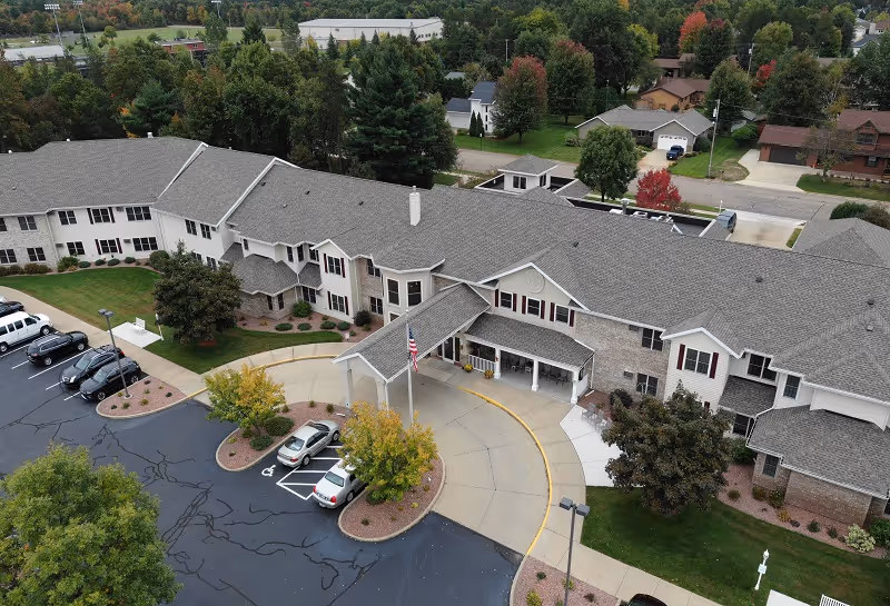 Aerial view of a large senior living facility building with a gray roof and beige exterior walls. The entrance has a covered driveway with an American flag on a pole. There are several cars parked in the parking lot, surrounded by landscaped areas with trees and bushes. The surrounding neighborhood has houses and trees with some fall foliage.