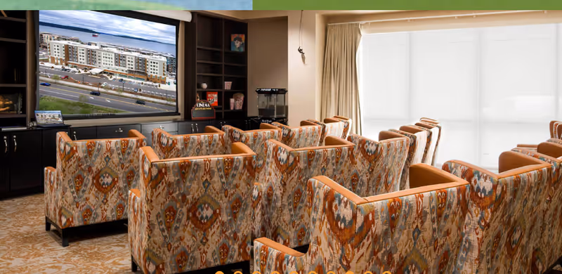 A cozy media room with multiple patterned armchairs arranged in rows facing a large flat-screen TV mounted on a dark wood cabinet. The TV displays an image of a building complex. The room has beige walls, a patterned carpet, and a large window covered with white blinds allowing natural light to enter.