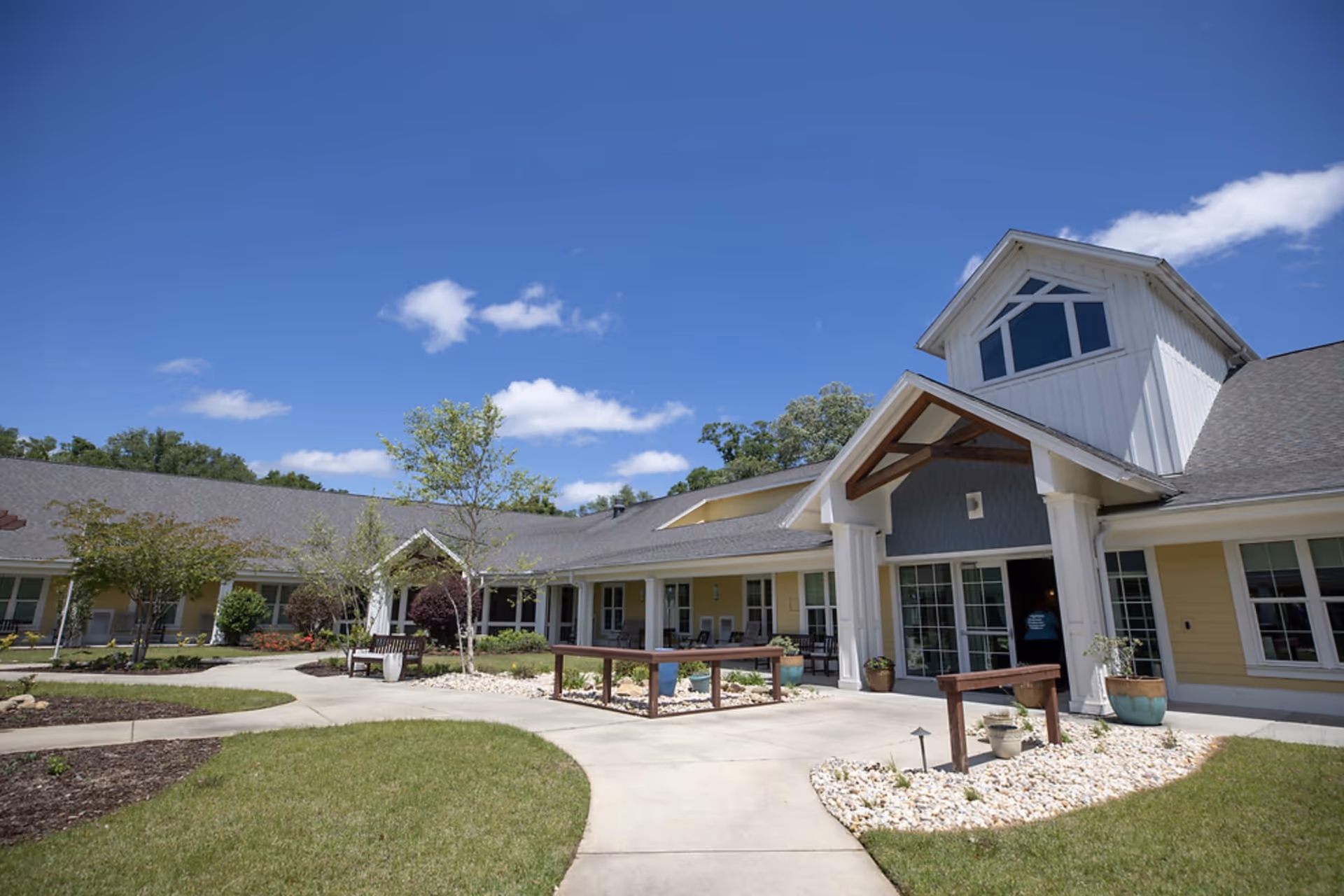 Exterior view of a senior living facility with a well-maintained garden, paved walkways, benches, and a building with large windows and a peaked roof under a clear blue sky.