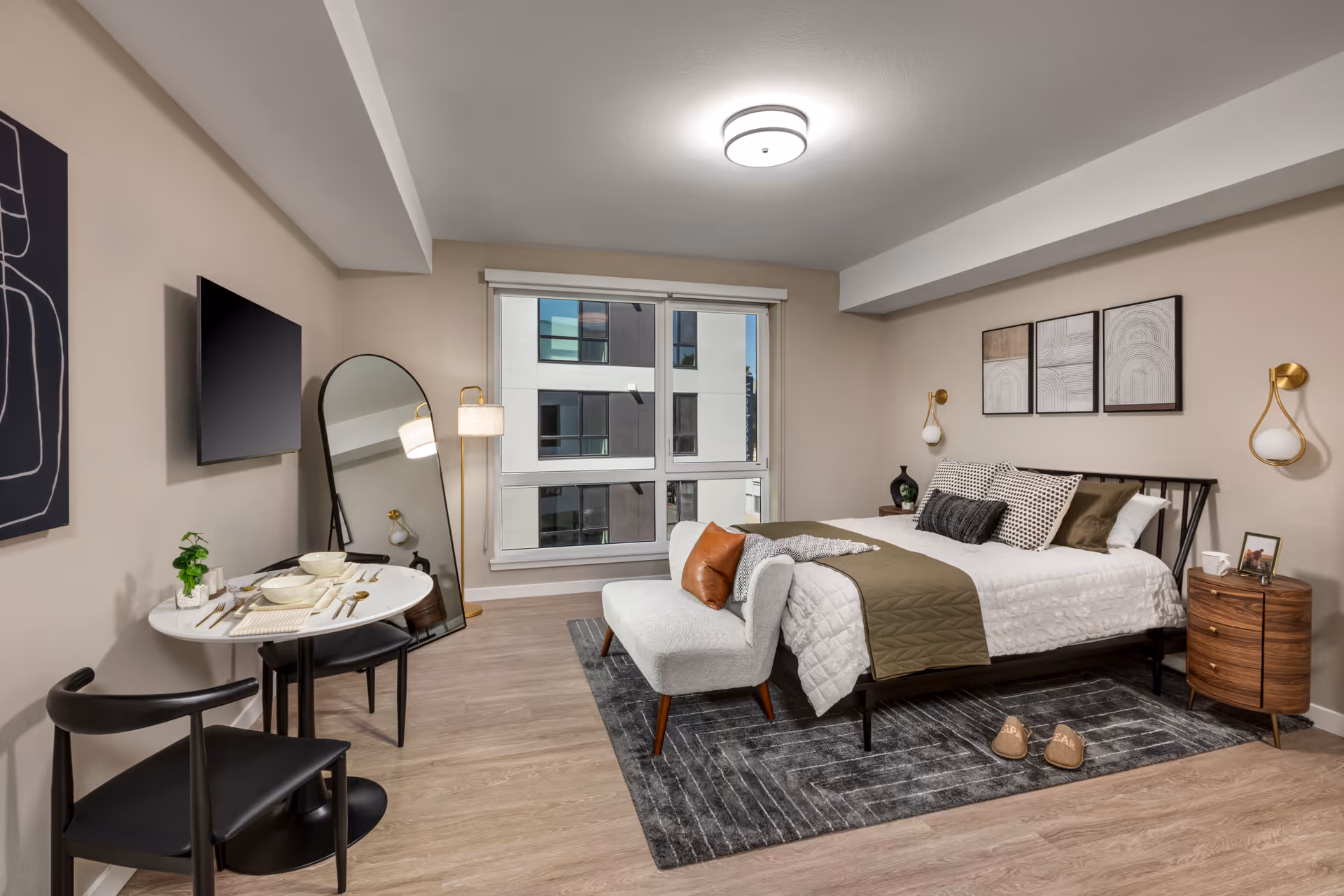 A modern bedroom in a senior living facility featuring a neatly made bed with multiple pillows and a green throw blanket. There is a small bench at the foot of the bed, a round wooden nightstand with a lamp and picture frame, and three framed artworks on the wall above the bed. A large window with a view of another building allows natural light into the room. On the left side, there is a small round dining table set for two with black chairs, a tall floor mirror, and a wall-mounted flat-screen TV. The room has light wood flooring and neutral-colored walls.