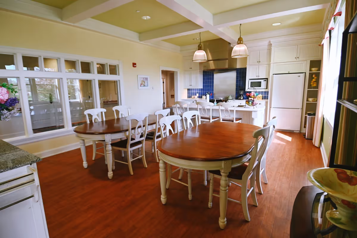 Bright communal dining room with wooden tables and white chairs adjacent to a kitchen with white cabinets and stainless appliances.