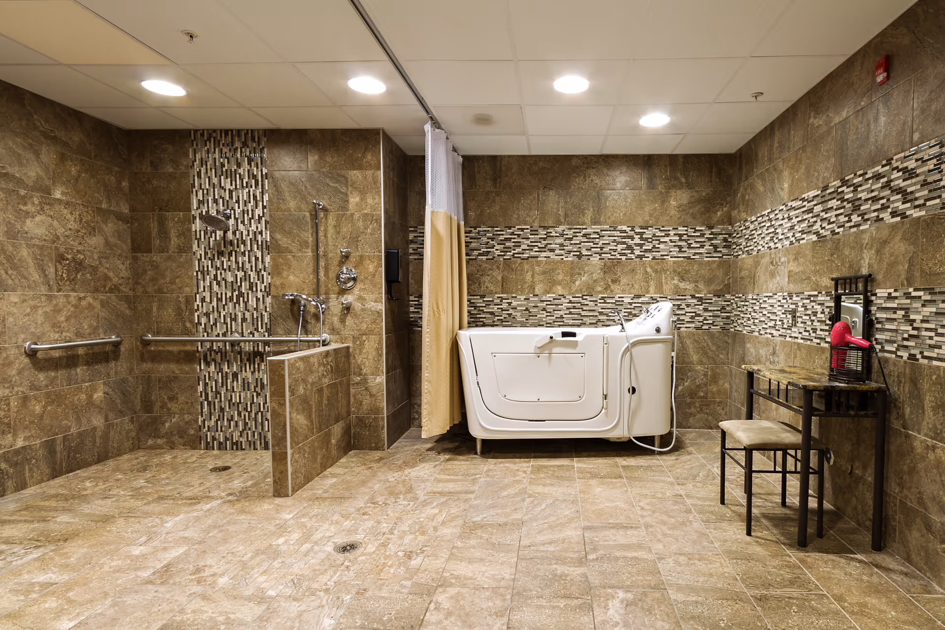 A spacious bathroom with brown tiled walls and floor, featuring a walk-in shower with grab bars and a handheld showerhead, a white walk-in bathtub with a door, and a small vanity table with a chair and a pink hairdryer on top.