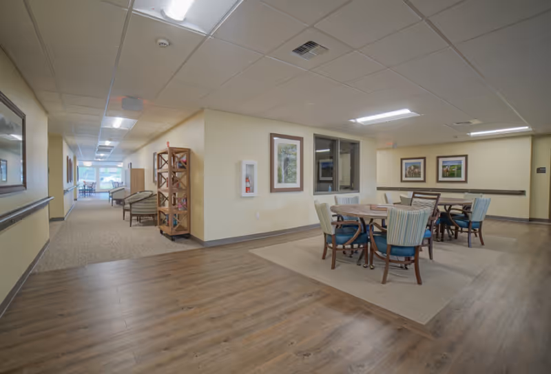 Interior common area of a senior living facility with round dining tables and chairs on a rug, wood-look floors, and a corridor with additional seating.