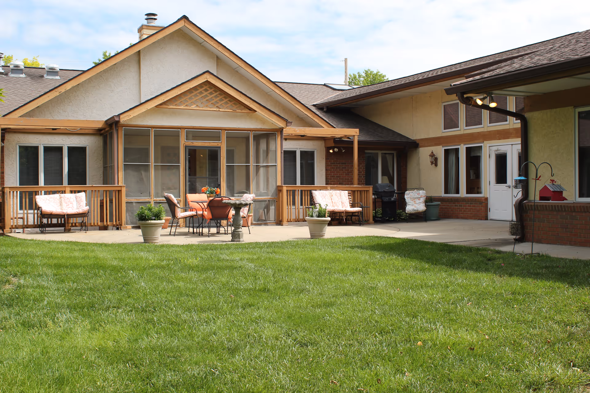 Exterior courtyard of a senior living building with a grassy lawn, patio seating, potted plants, and a screened porch.