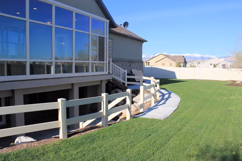 A backyard area of a residential building with a green lawn, a white fence, a concrete pathway, and a large windowed sunroom attached to the house. In the background, there are other houses and snow-capped mountains under a clear blue sky.