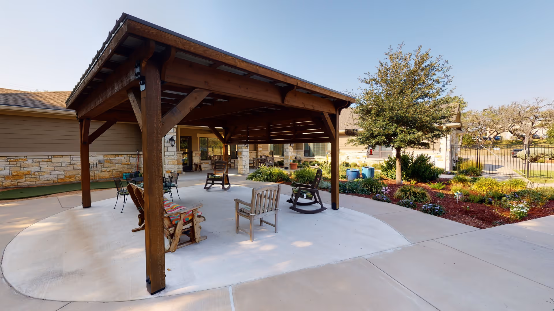 Outdoor seating area at Mercy House Living Springs featuring a wooden pergola with several chairs underneath, surrounded by a concrete patio and landscaped garden with trees and shrubs.