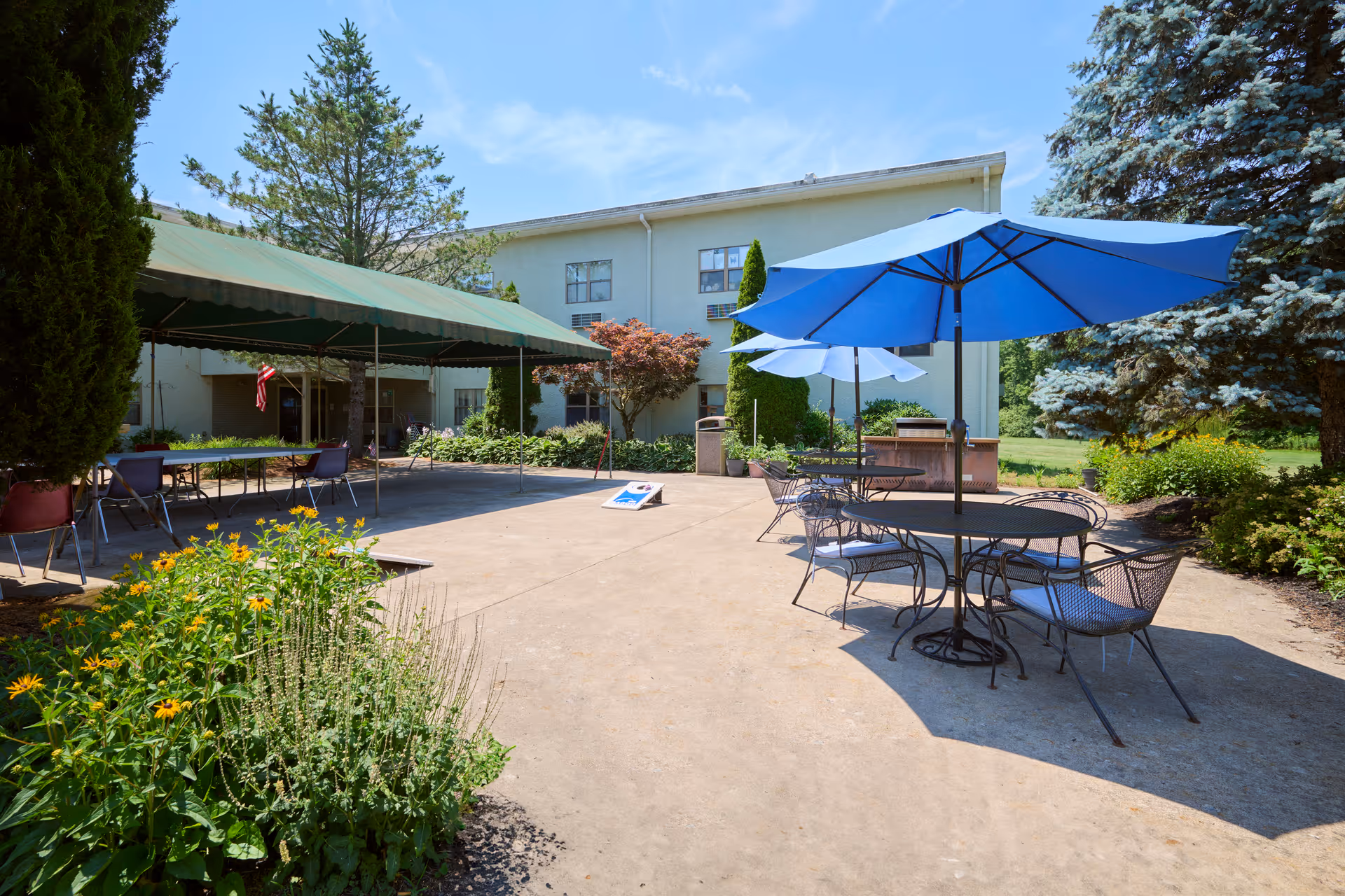 Outdoor patio area at Birchwood Rehabilitation and Healthcare Center with tables and chairs under blue umbrellas, a green canopy covering additional seating, surrounded by trees and plants under a clear blue sky.