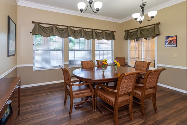 A dining room with a round wooden table surrounded by six cushioned chairs. The room has large windows with white blinds and green valances. There are two small flower arrangements on the table, wooden flooring, beige walls with white trim, and two ceiling light fixtures.