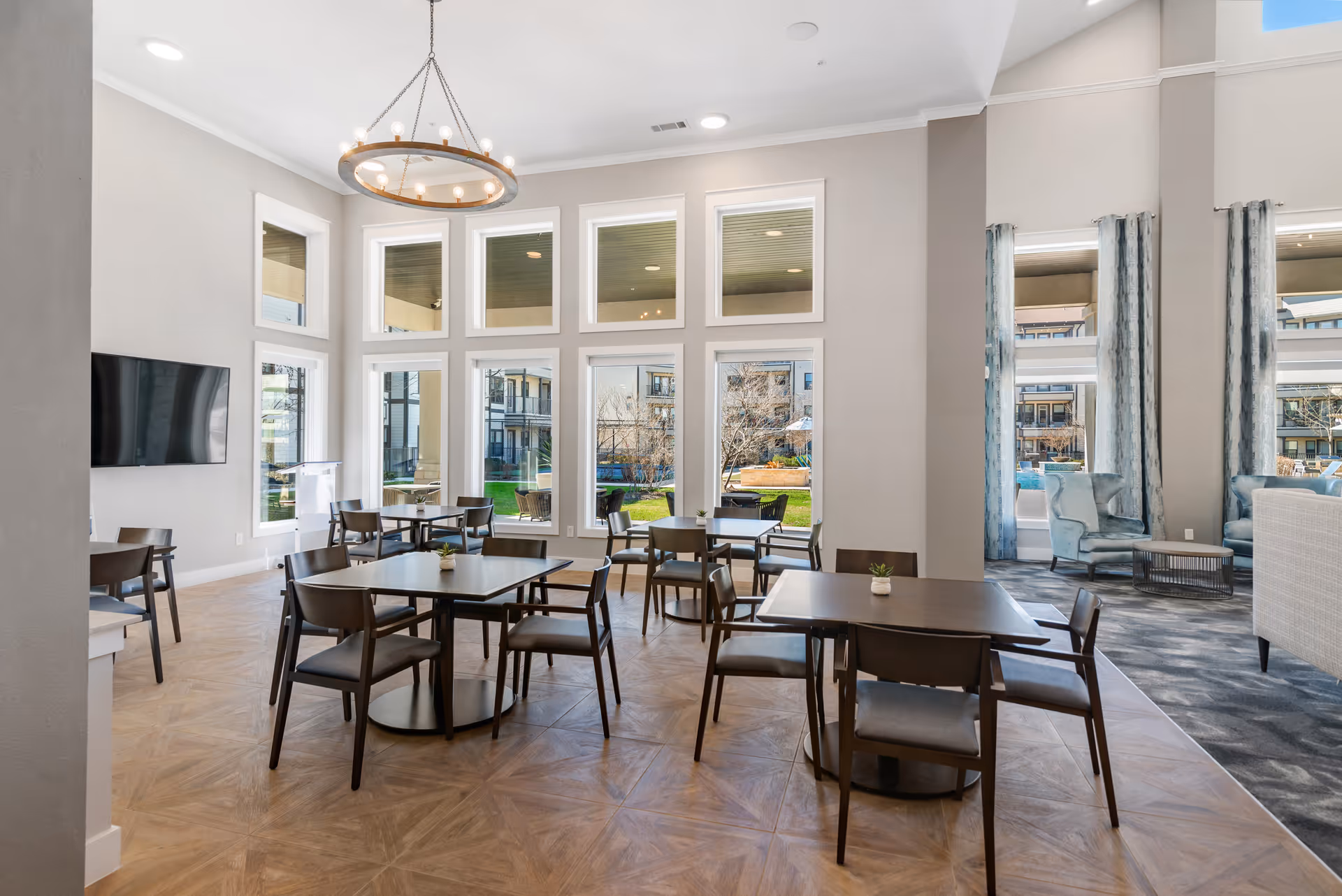 Bright communal dining and lounge area with several tables and chairs, large floor-to-ceiling windows overlooking a courtyard, a chandelier, and a wall-mounted TV.