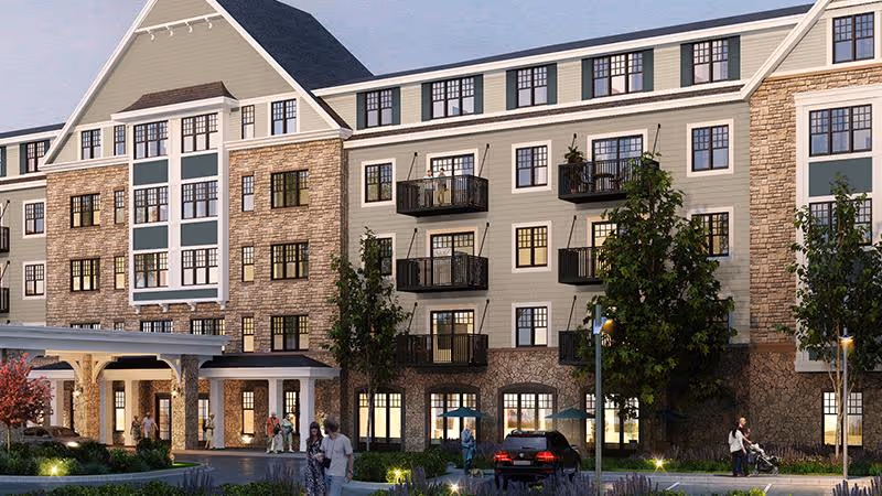 Front exterior of a multi-story senior living building with balconies, an entrance canopy, landscaping, parked cars, and people near the driveway.