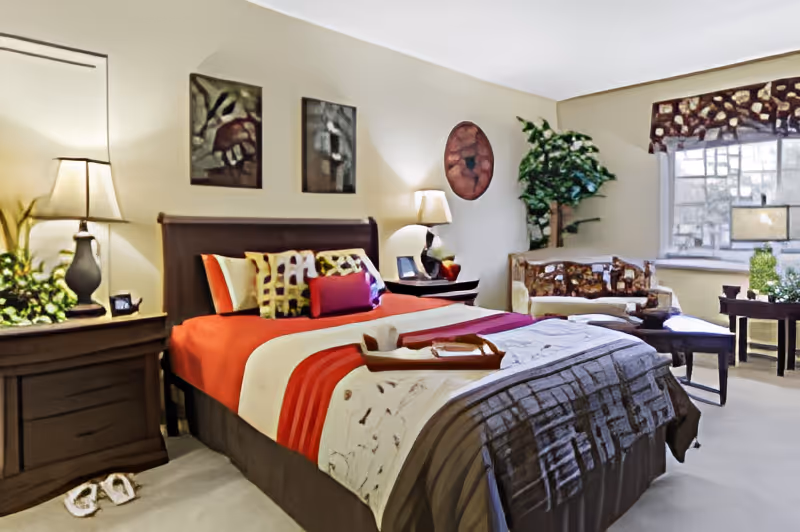 A cozy bedroom with a large bed dressed in red, white, and brown bedding. The bed has multiple decorative pillows and a wooden tray on top. On either side of the bed are nightstands with lamps and small plants. There is a patterned loveseat near a window with a valance, and a small table with plants beside it. The walls have several pieces of artwork, and the room is softly lit, creating a warm and inviting atmosphere.