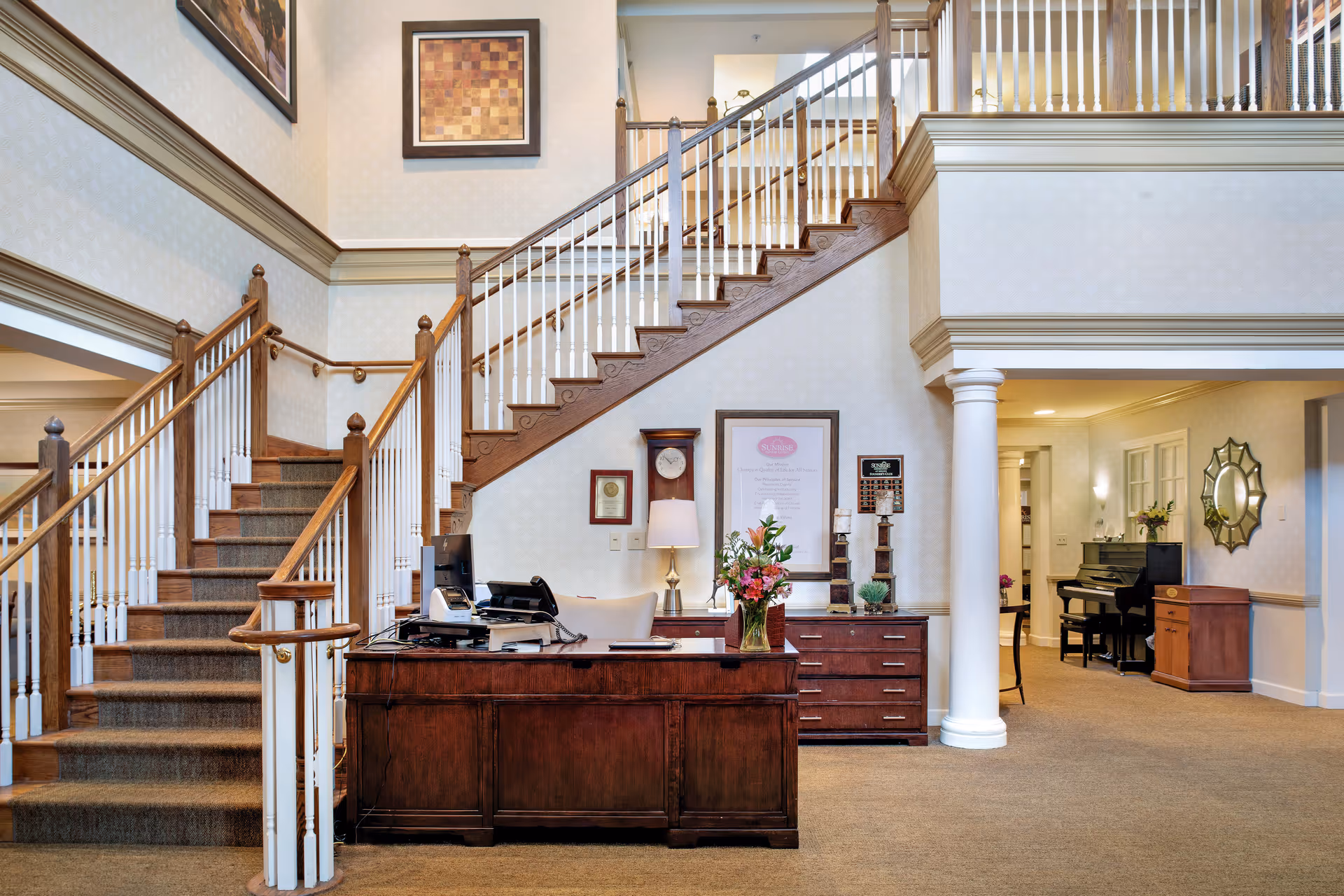 Interior view of a senior living facility reception area with a wooden desk, a computer, a telephone, and a vase of flowers. Behind the desk is a staircase with wooden steps and white railings leading to an upper floor. The walls are decorated with framed artwork and a clock. To the right, there is a hallway with a piano, a mirror, and additional furniture.