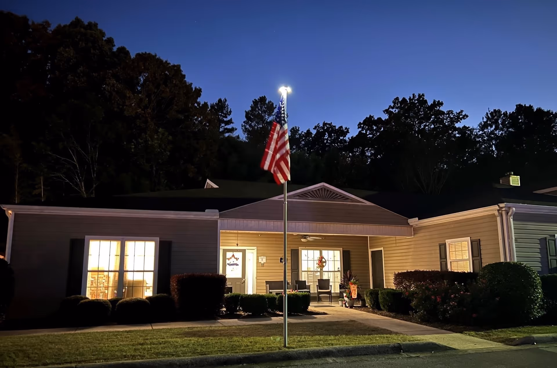 Exterior view of a single-story building at dusk with an American flag on a flagpole in the foreground. The building has lit windows and a covered entrance with chairs and some decorations. Trees are visible in the background under a darkening sky.