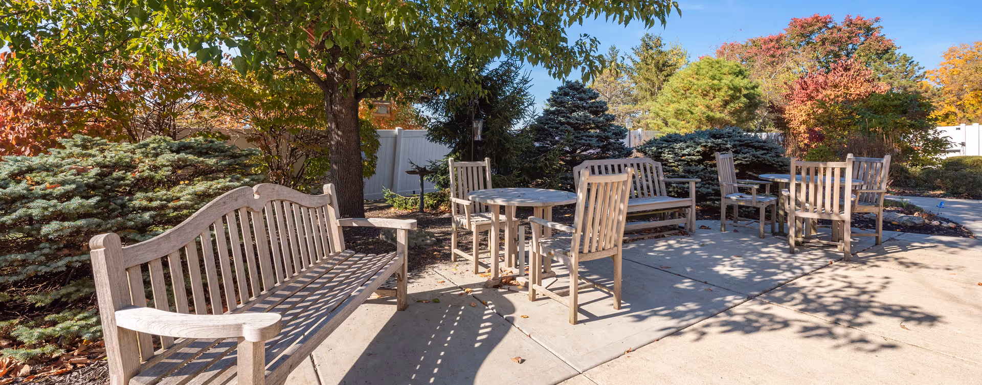 Sunlit outdoor seating area with wooden benches and chairs surrounded by trees and shrubs.