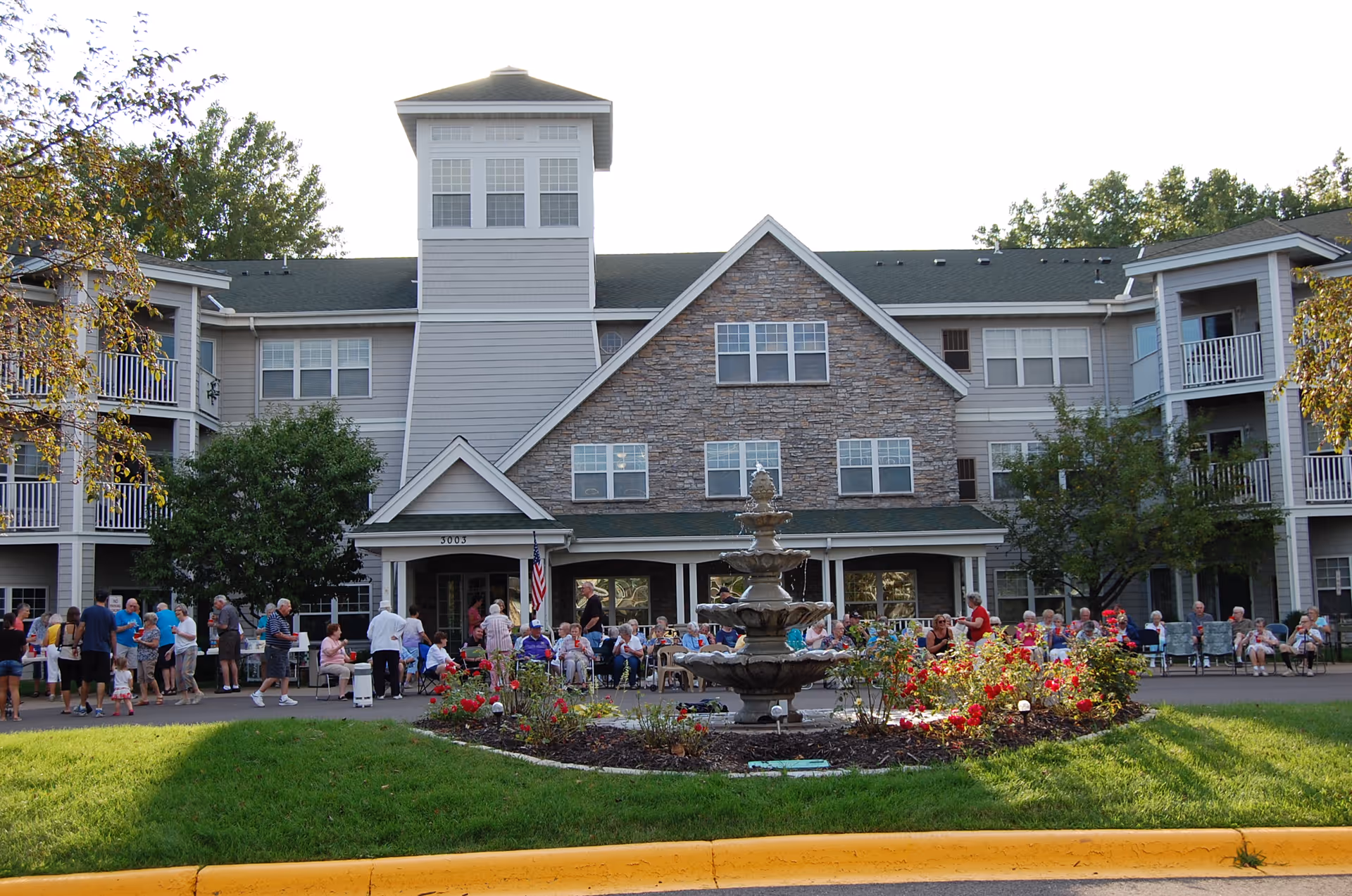 Front exterior view of a senior living facility named Cardinal Pointe of Maplewood with a large group of elderly people and visitors gathered outside near a multi-tiered fountain surrounded by flower beds. The building has multiple windows, balconies, and a stone facade in the center section.