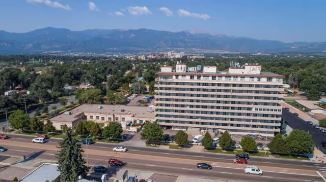 Aerial view of a multi-story post-acute rehabilitation building beside a road with cars, trees, and distant mountains.