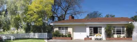 Exterior of a single-story assisted living facility with white walls, a red roof, and front shrubs.