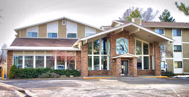 A brick senior living facility with large glass entrance and sloped roof, parking lot in foreground.