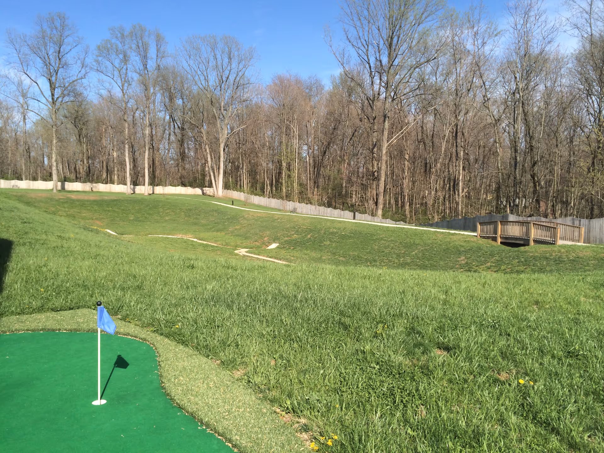 A grassy outdoor area with a small putting green and blue flag in the foreground, rolling lawn, a wooden bridge, fence, and leafless trees in the background.