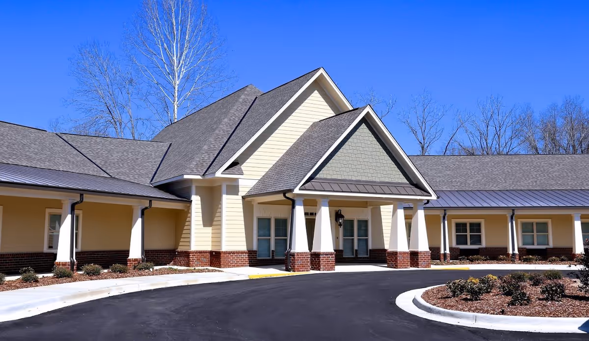 Front entrance of a single-story senior living facility with a covered drive-up, white columns, brick accents, and a landscaped circular driveway under a clear blue sky.