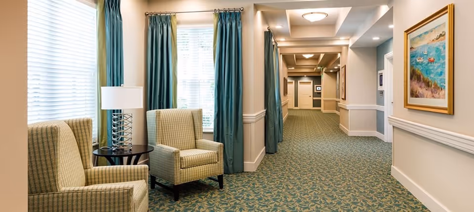 A well-lit hallway in a senior living facility with patterned carpet and beige walls. Two upholstered armchairs with a small round table and a lamp are placed near large windows with teal curtains. The hallway features framed artwork on the walls and multiple doors along the corridor.