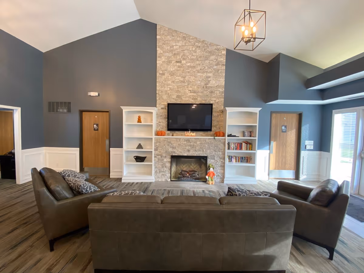 A cozy senior living facility common area with a stone fireplace centered between two white bookshelves. Above the fireplace is a mounted flat-screen TV. The room features gray walls with white wainscoting, a high vaulted ceiling with a modern light fixture, and wood-look flooring. There are three brown leather sofas arranged facing the fireplace. Two wooden doors with restroom signs are visible on either side of the fireplace, and large windows on the right let in natural light.