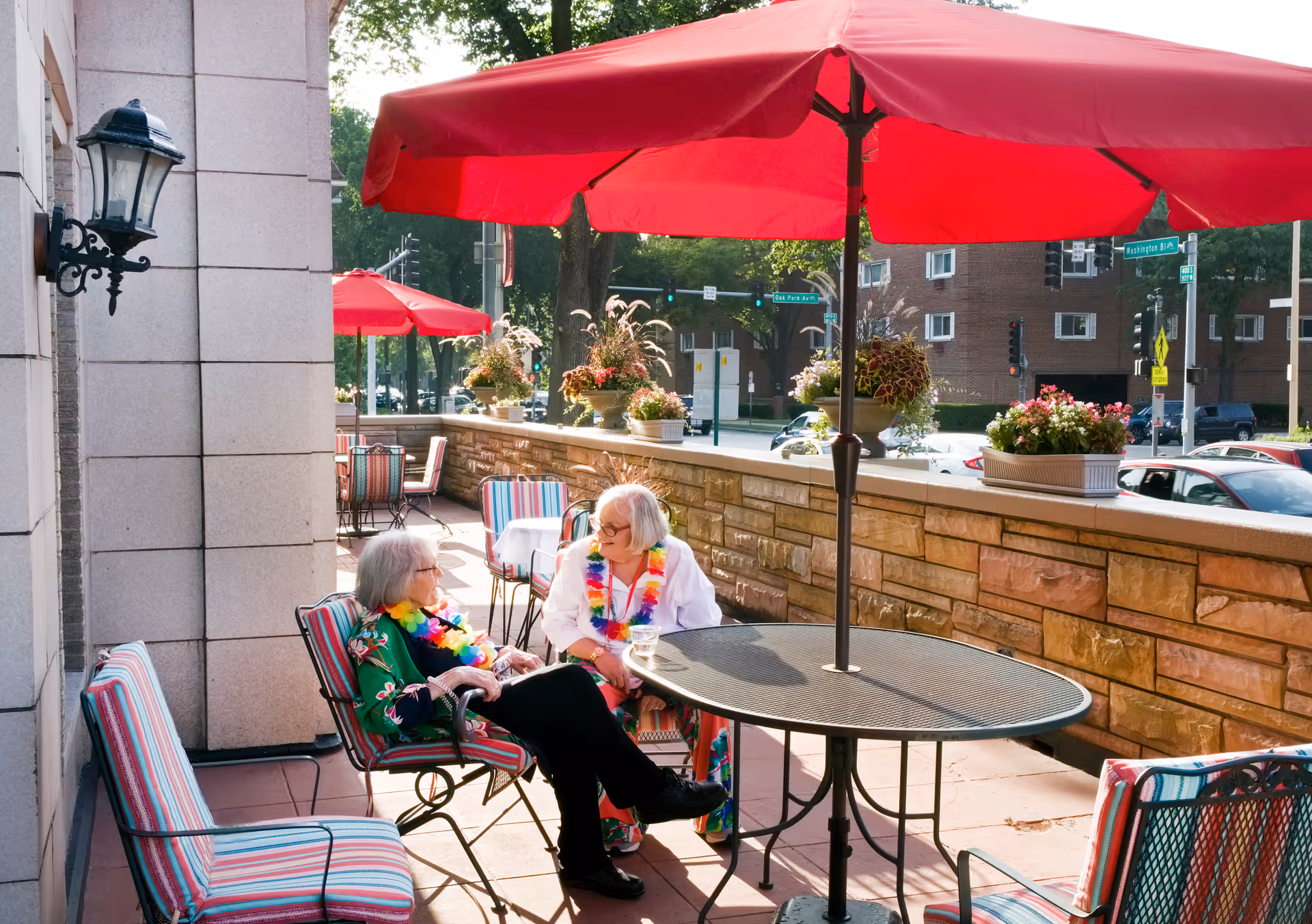 Two elderly women sitting and chatting at a round outdoor table with a large red umbrella on a patio. The patio has striped cushioned chairs, potted plants on a stone wall, and a street with cars and buildings in the background.