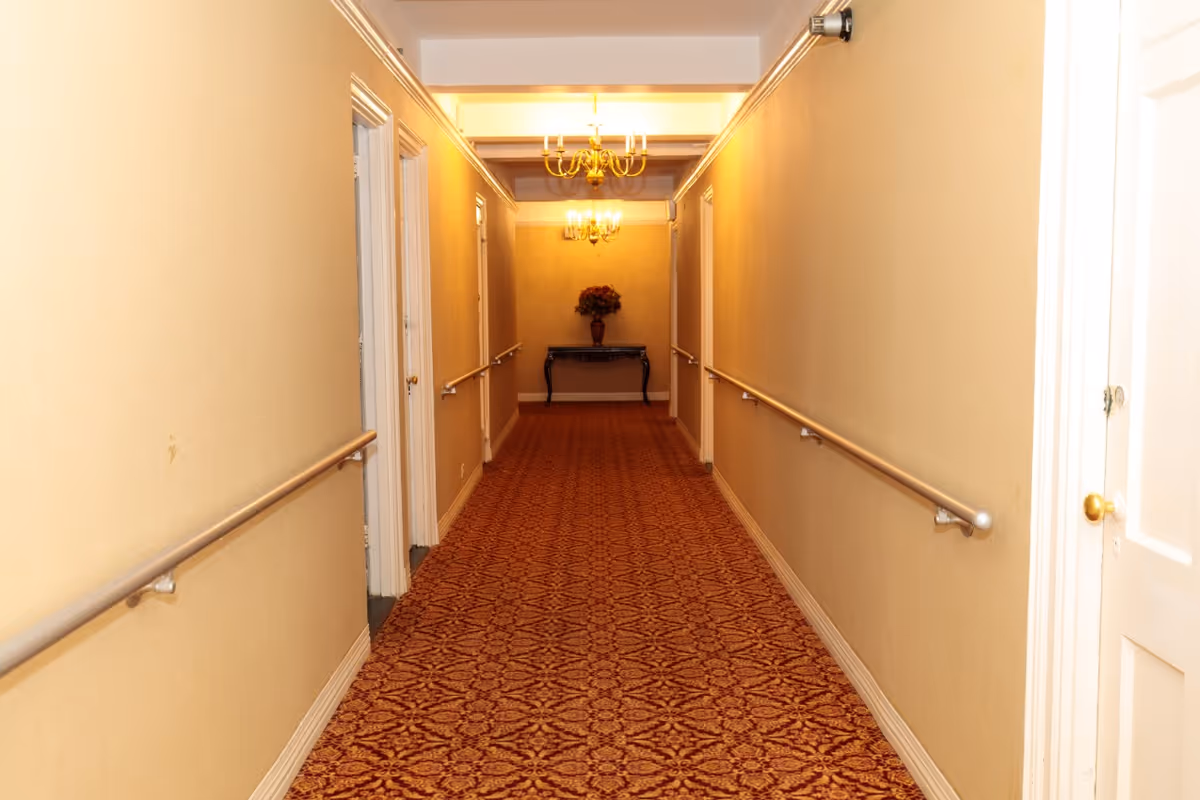 A long, narrow hallway with beige walls and a patterned red carpet. There are handrails on both sides of the hallway and several white doors along the walls. At the end of the hallway, there is a small table with a vase of flowers and two chandeliers hanging from the ceiling providing warm lighting.