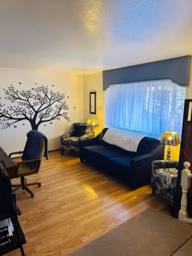 A cozy living room with wooden flooring, featuring a dark blue sofa with a white throw blanket, two patterned armchairs, a desk chair, and two table lamps with patterned lampshades. A large window with sheer white curtains and a gray valance lets in natural light. A decorative black tree wall decal with birds is on the left wall.